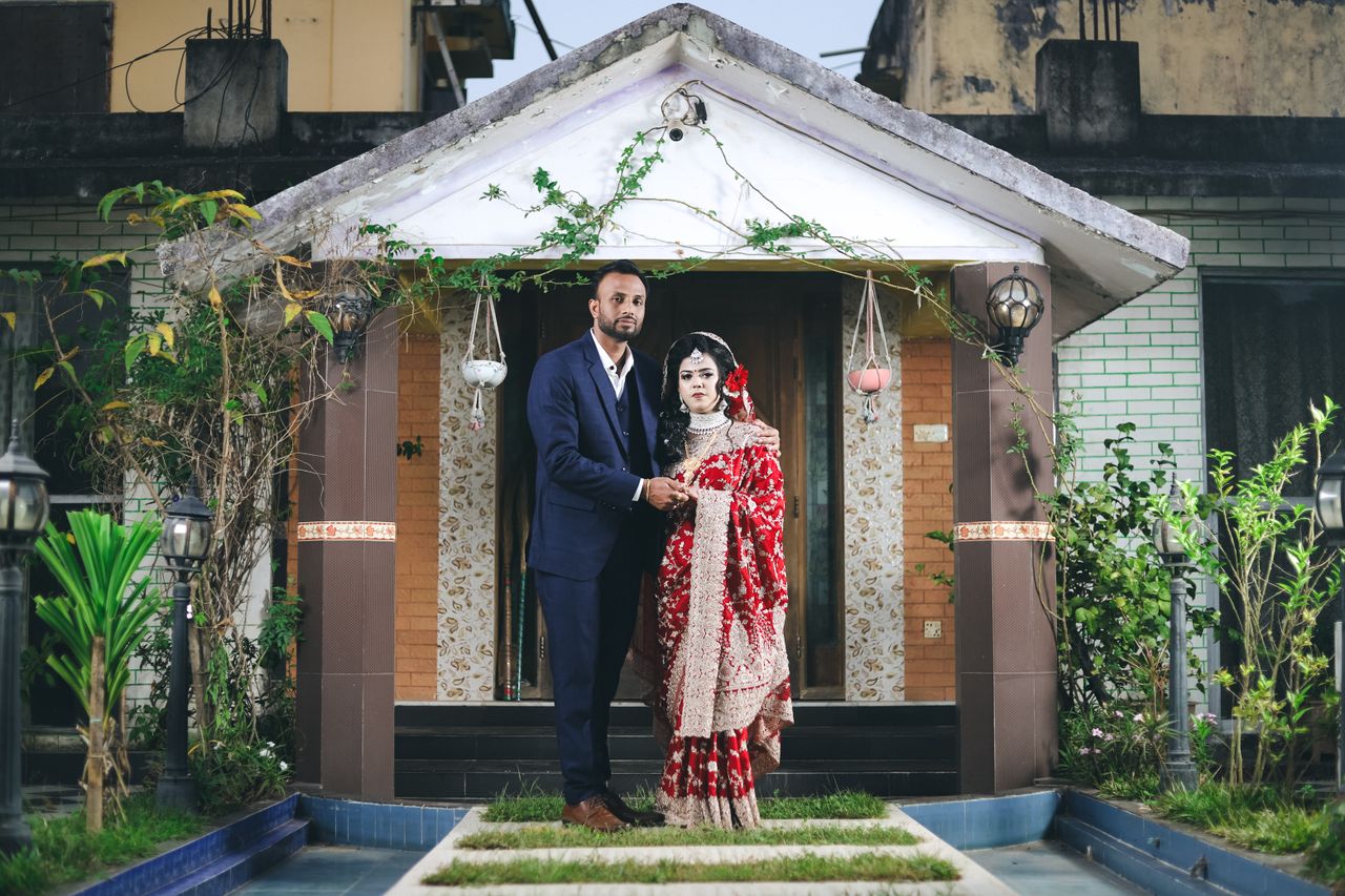 Indian couple in traditional wedding attire posing outside a decorated house